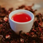 فنجان سفید حاوی چای ترش قرمز روی گل های خشک شده هیبیسکوس